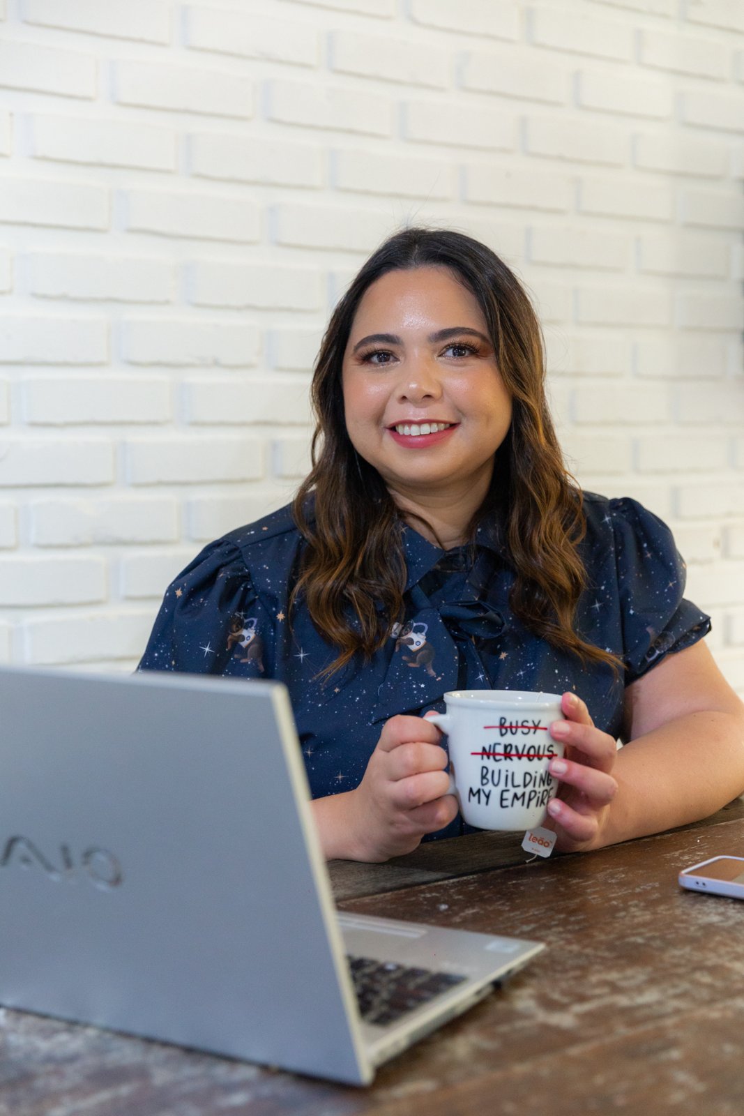 Mayra Pires sentada à mesa com o notebook, sorrindo